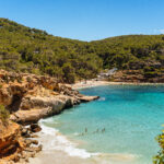 View of Cala Salada from Cala Saladetta with turquoise waters and rocky coastline in Ibiza.